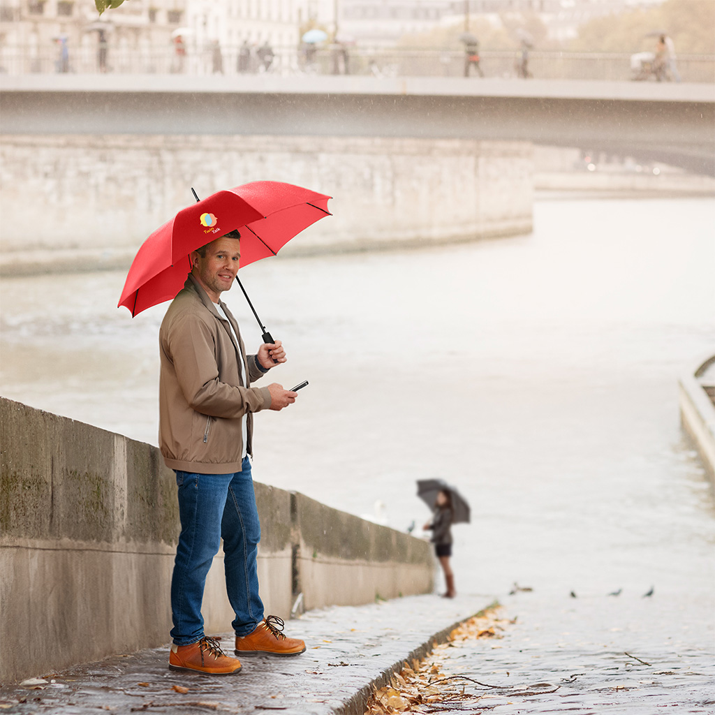 Mann mit FARE® AC-Stockschirm und Handy in der Hand an Flussufer