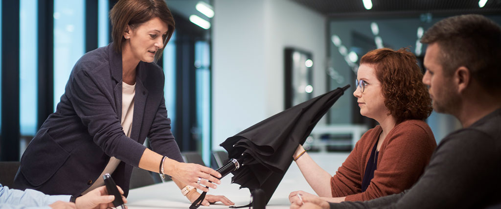 Beratungsszene im Büro: Eine Mitarbeiterin zeigt am Tisch einem Team einen geöffneten Regenschirm und erklärt Details.