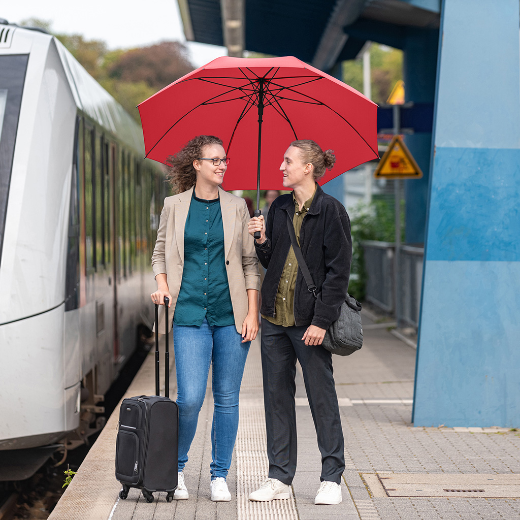 Zwei Personen stehen lächelnd an einem Bahnsteig unter einem großen, roten Gästeschirm. Links steht eine Frau mit Brille, beigem Sakko und einem schwarzen Rollkoffer; rechts eine Person mit Dutt, dunkler Jacke und Umhängetasche, die den Schirm hält. Im Hintergrund ist ein weißer Regionalzug eingefahren.