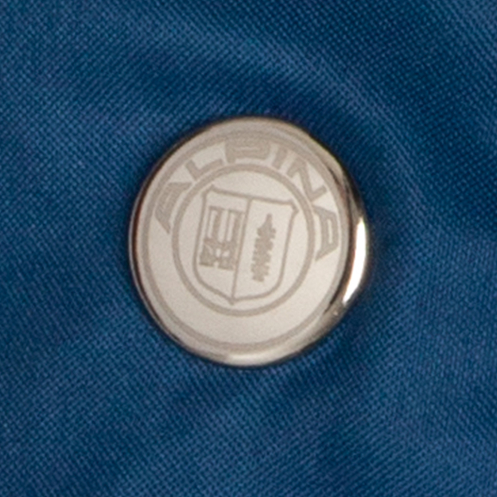 Nahaufnahme eines silberfarbenen Druckknopfs auf blauem Schirmstoff. Auf dem runden Knopf ist ein individuell gestaltetes Emblem mit Schriftzug und Wappensymbol zu sehen.