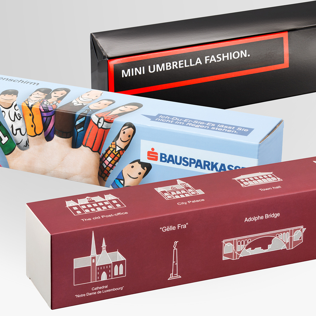 Drei individuell gestaltete Schirmverpackungen in unterschiedlichen Designs. Zu sehen sind eine schwarze Verpackung mit „MINI UMBRELLA FASHION.“, eine hellblaue Verpackung mit Bausparkasse-Motiv und eine bordeauxrote Verpackung mit weißen Illustrationen von Sehenswürdigkeiten.