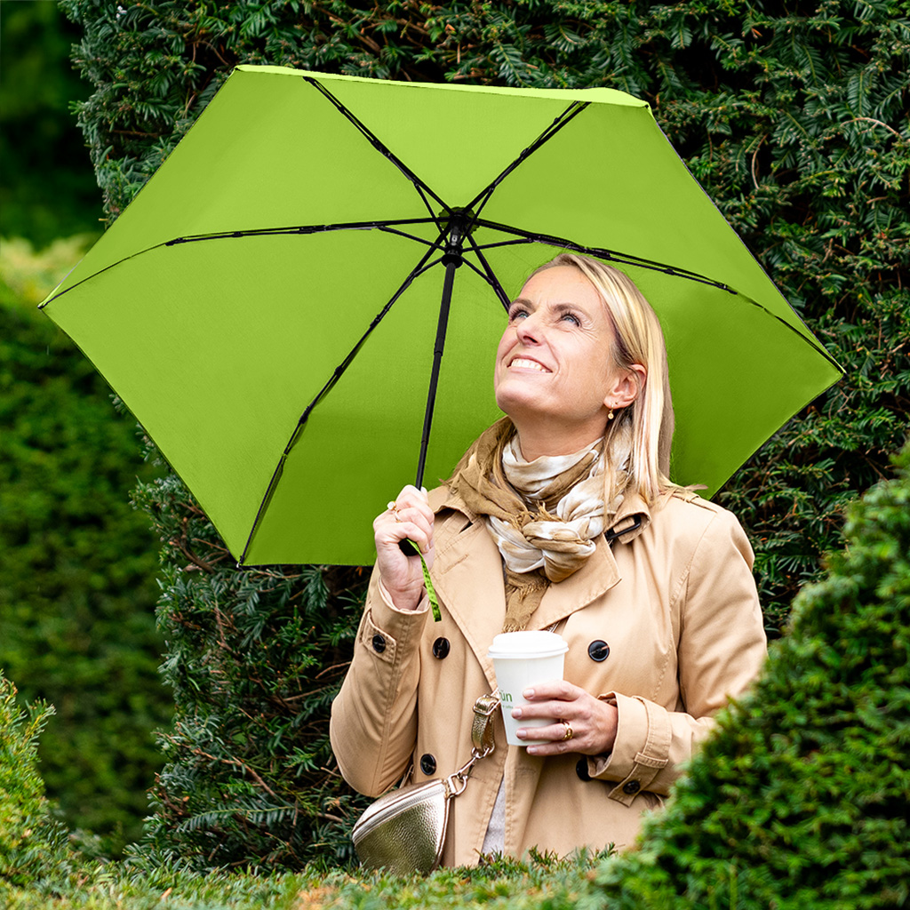 Frau mit Regenschirm und Kaffebecher hinter einer gründen Hecke guckt in den Himmel