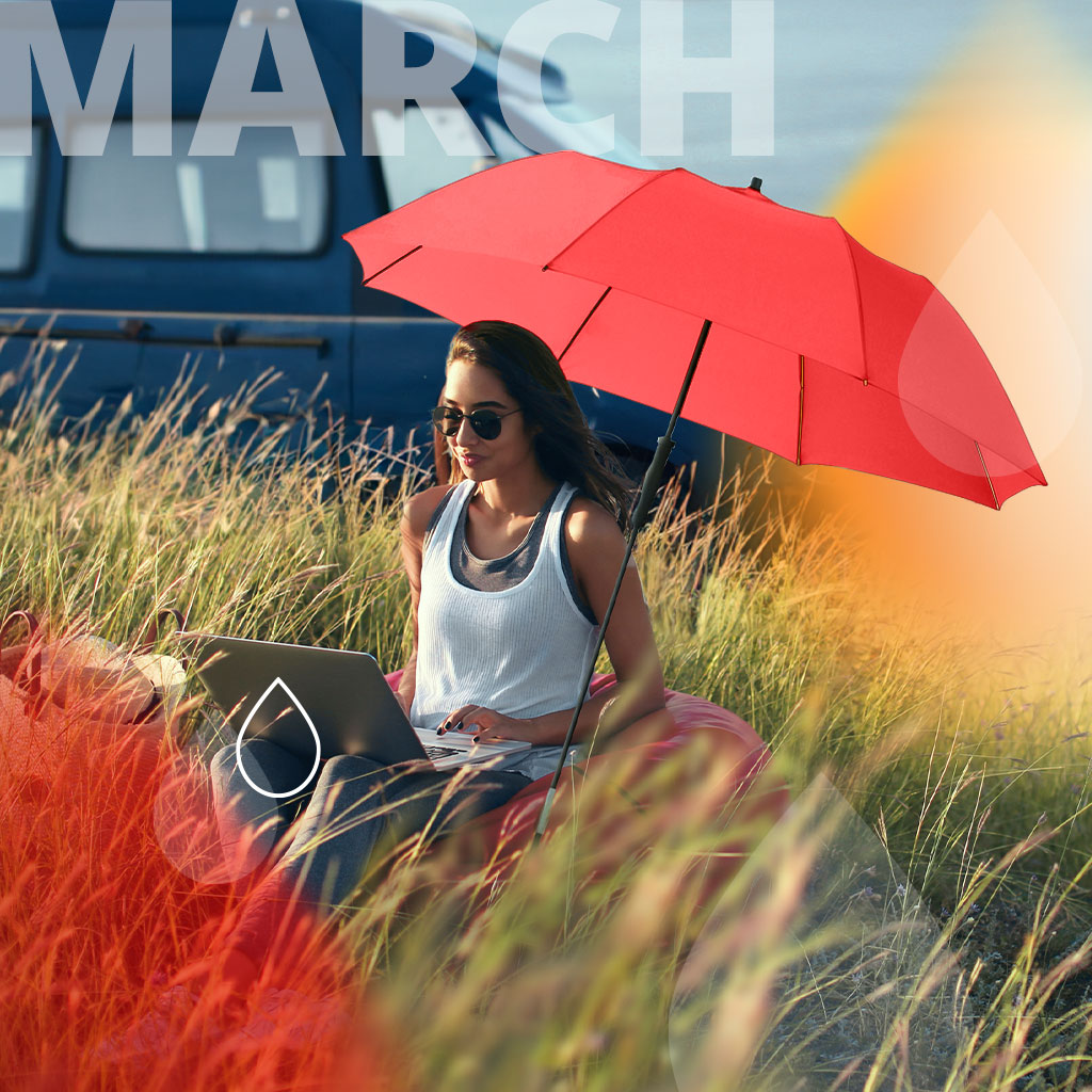 Umbrella of the Month March: Woman with red parasol working on laptop on a hill in the sun. Text: March