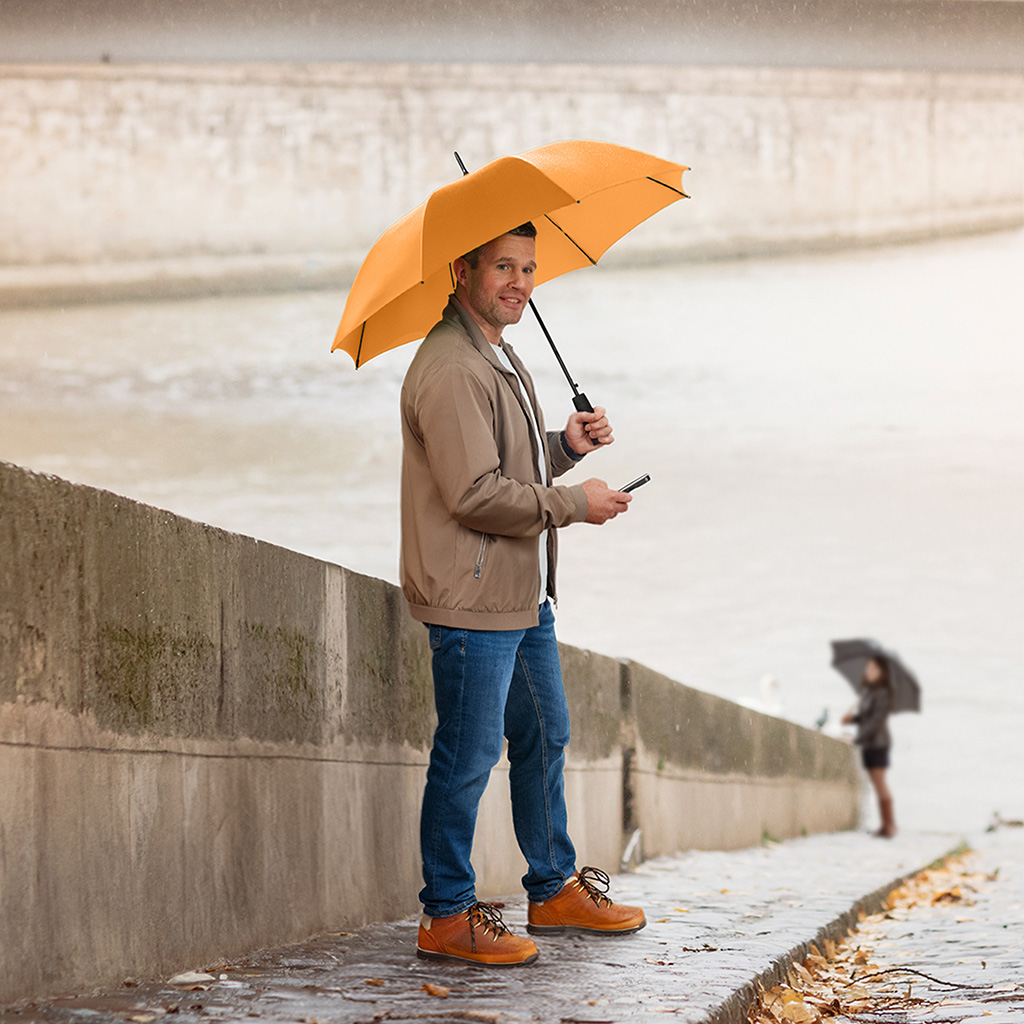 Ein Mann steht an einem regnerischen Tag mit einem leuchtend gelben Stockschirm auf einem gepflasterten Uferweg an einem Fluss. Er trägt eine beige Bomberjacke, Jeans und braune Lederschuhe und blickt lächelnd in die Kamera, während er ein Smartphone in der Hand hält. Im Hintergrund ist eine weitere Person mit einem dunklen Schirm sowie eine Steinmauer zu sehen.