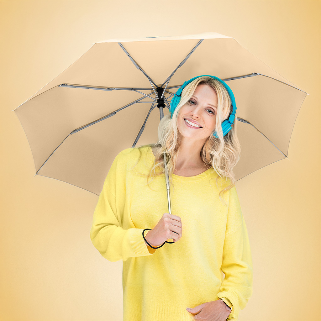 Eine lächelnde junge Frau mit blondem Haar trägt einen hellgelben Pullover und auffällige türkisfarbene Kopfhörer. Sie hält einen aufgespannten, beigefarbenen Taschenschirm über sich. Das Bild ist vor einem schlichten, passend gelblich-beigen Hintergrund aufgenommen.