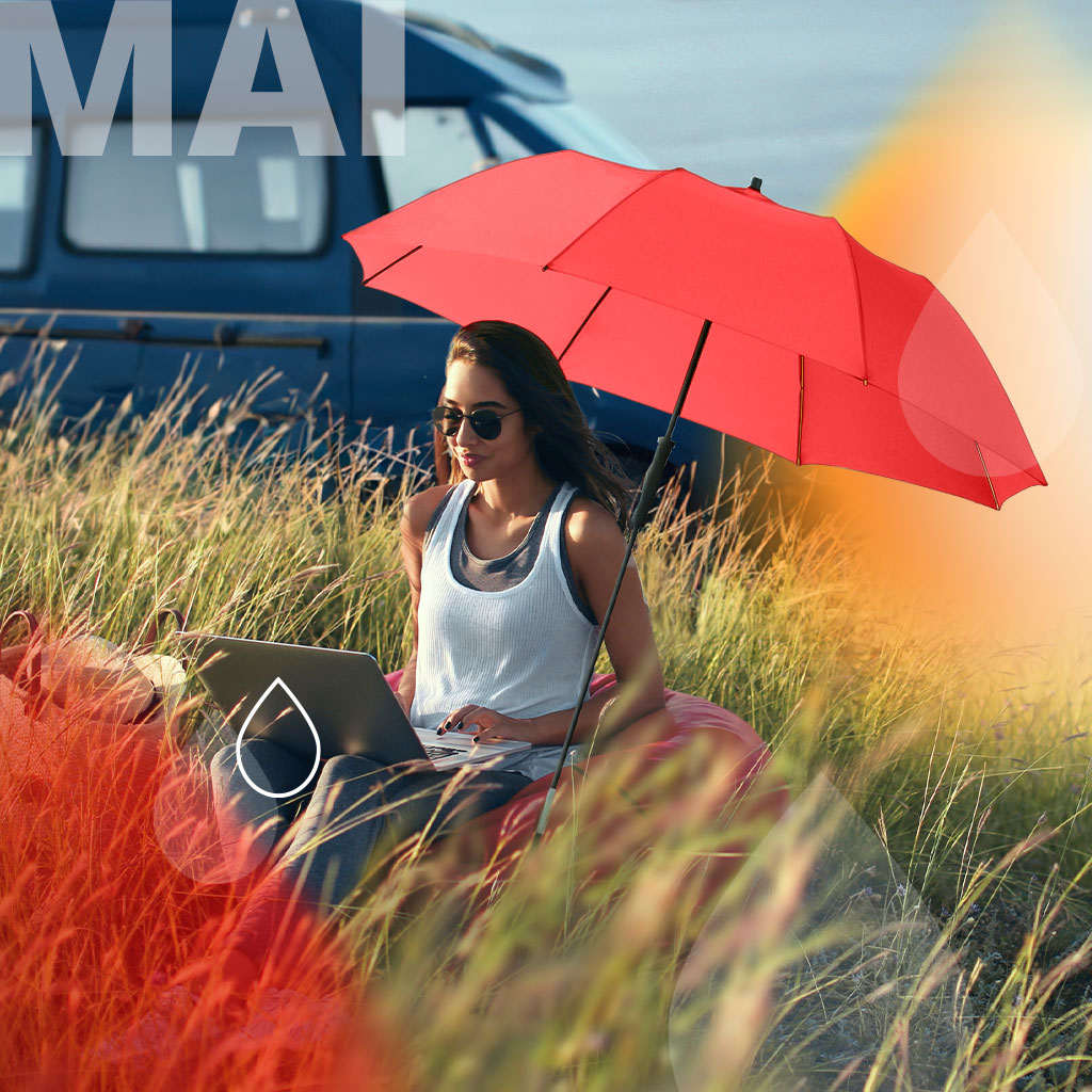 Schirm des Monats Mai: Frau an in den Dünen sitzend unter dem Sonnenschirm Travelmate Camper in rot.. Text: Mai