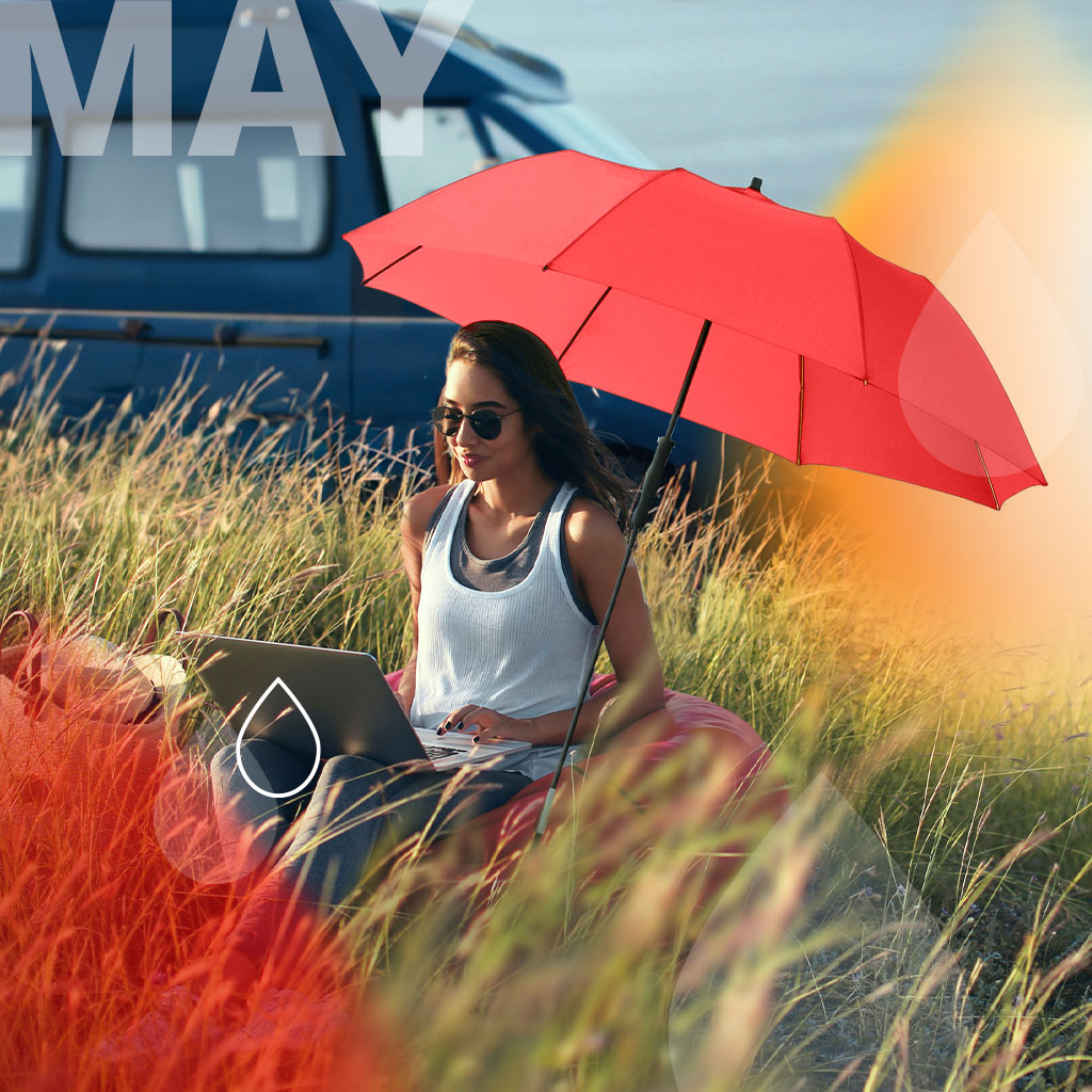 Umbrella of the month May: Woman sitting in the dunes under a parasol Travelmate Camper in red. Text: May